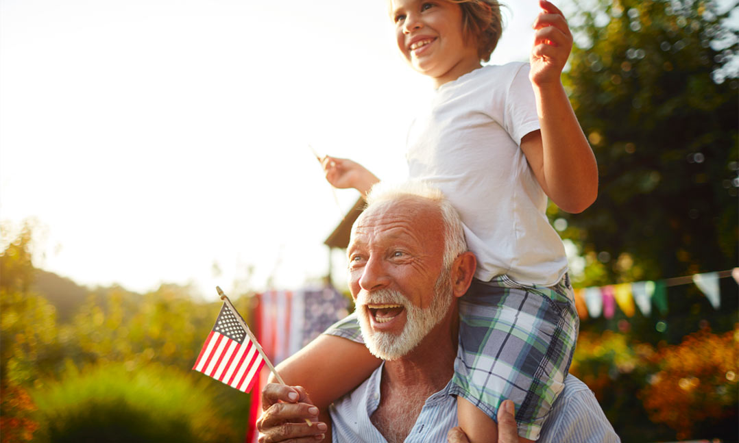 Older grandpa with white beard holding grandson on his shoulder while holding US flags